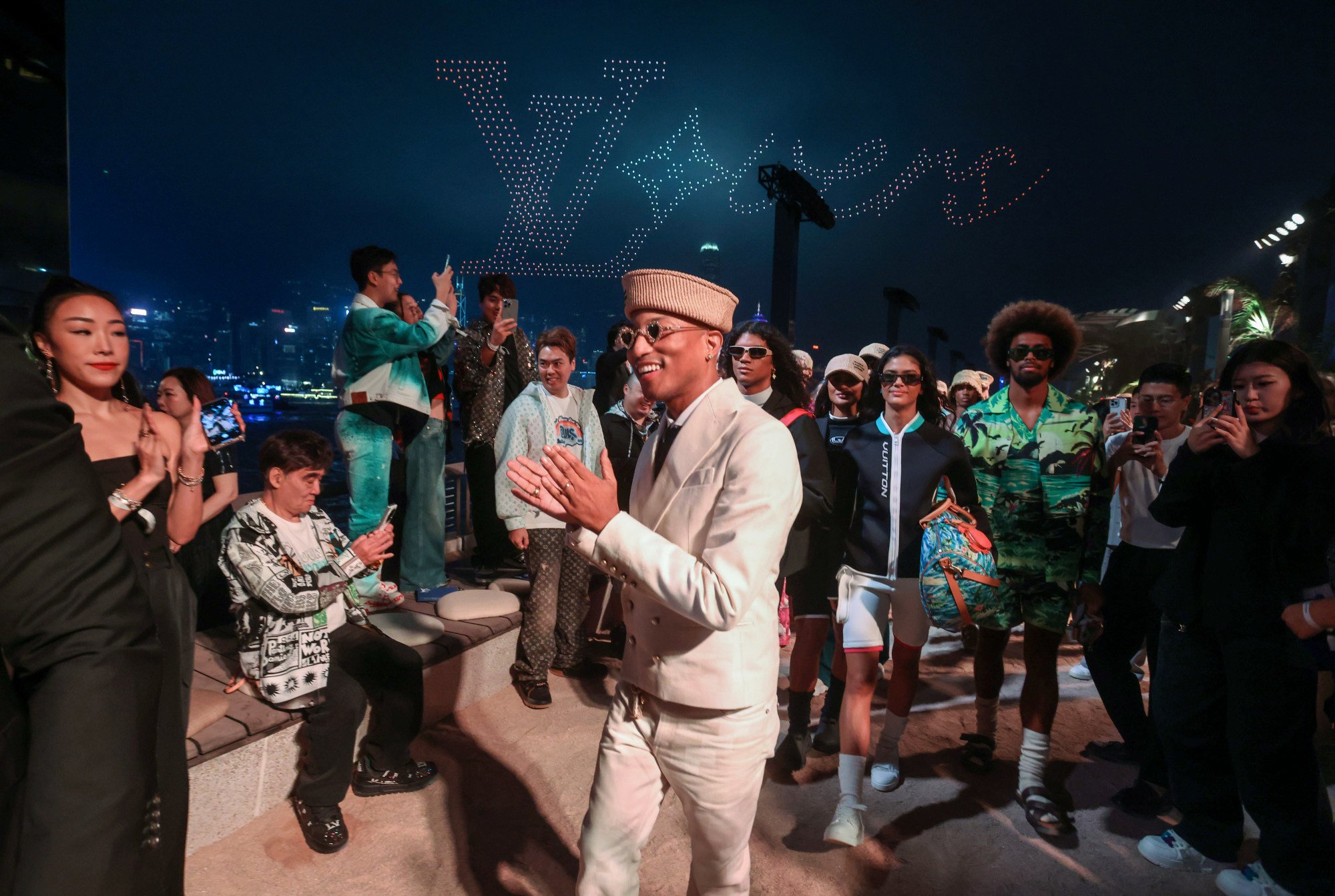 Giám đốc sáng tạo thời trang nam Louis Vuitton Pharrell Williams (giữa đeo kính) với các người mẫu sau lưng và nền trời là drone xếp hình biểu tượng LV tại buổi diễn. Ảnh: Jonathan Wong